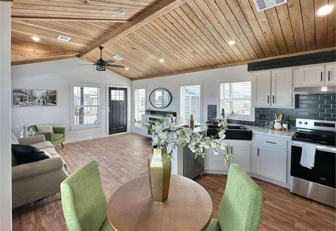 Modern open-concept living space with wood-plank ceiling, featuring a kitchen with black tile backsplash, dining area, and cozy seating. Bright and inviting.
