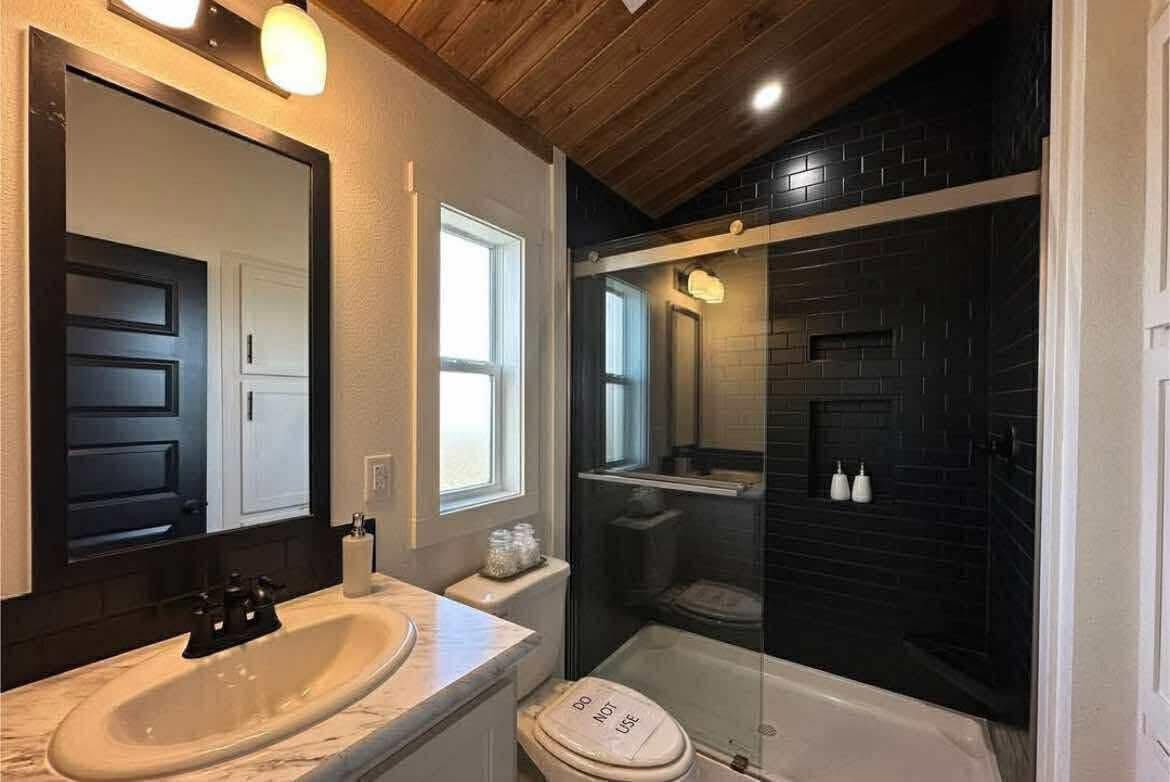 Modern bathroom with wooden ceiling and black tiled shower. Features a white sink, mirror, and toilet with “Do Not Use” sign. Bright and clean ambiance.