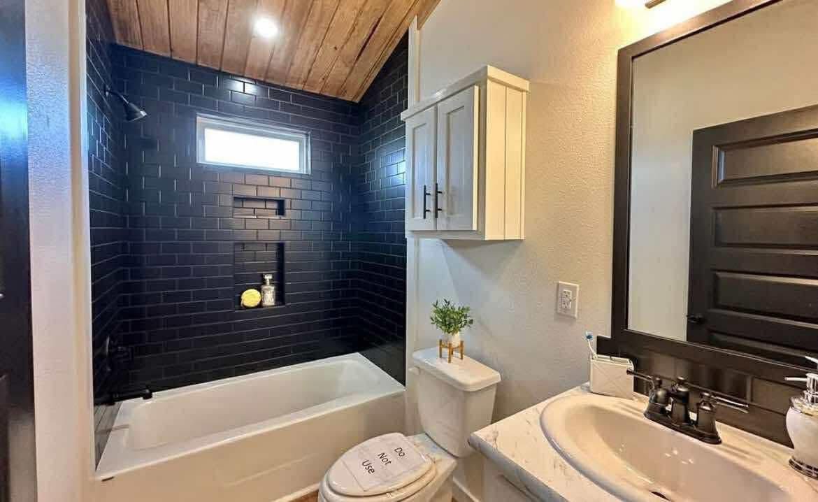 A modern bathroom with a black subway tile shower, white bathtub, and wooden ceiling. White vanity with sink, black-framed mirror, and decorative plant.