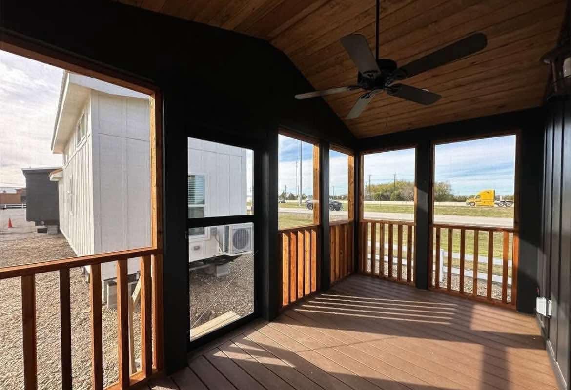 A cozy screened porch with wooden flooring and walls, featuring a ceiling fan. Sunlight filters in, casting shadows. View of a white building and road outside.