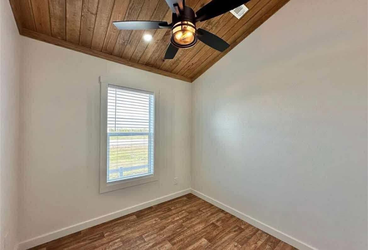 Small room with white walls, a wooden ceiling, and a matching wood floor. A window with blinds lets in natural light, and a modern black ceiling fan hangs overhead.