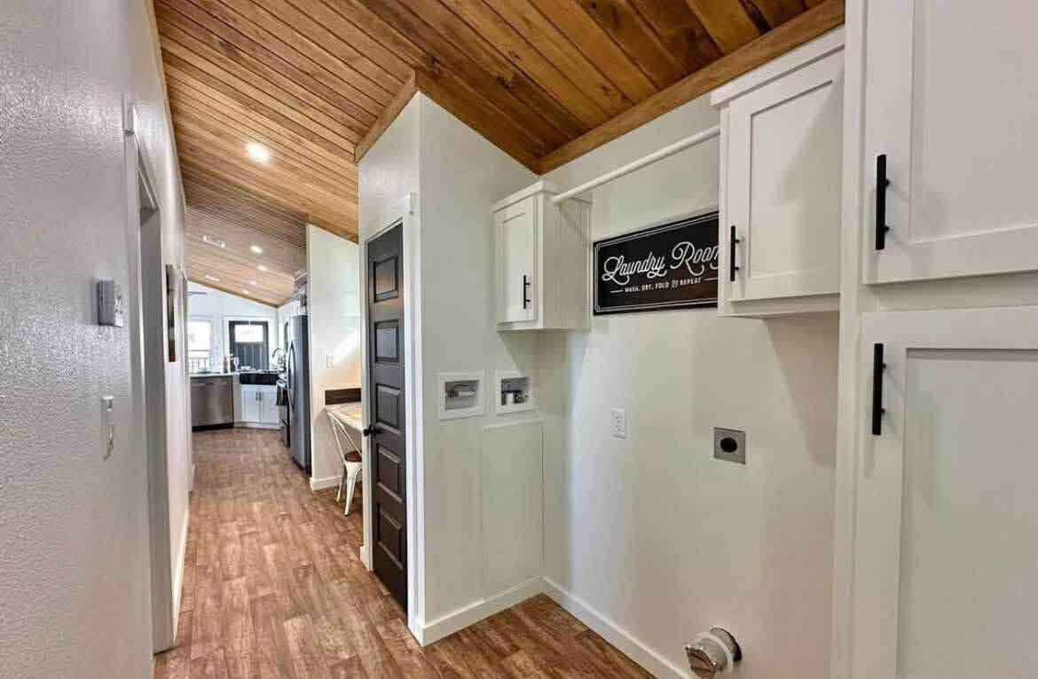 Hallway with wooden floor and ceiling, white walls, and cabinetry on the right with "Laundry Room" sign. Bright, modern, and inviting space.