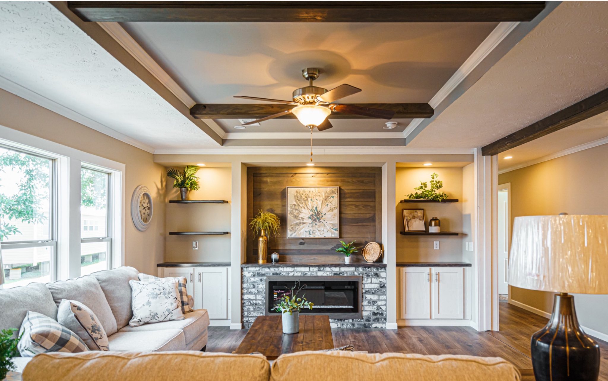A cozy living room with a modern ceiling fan and warm lighting. The space features a beige sofa, a wooden coffee table, and built-in shelves around a fireplace. Walls are light-colored, creating a welcoming atmosphere.
