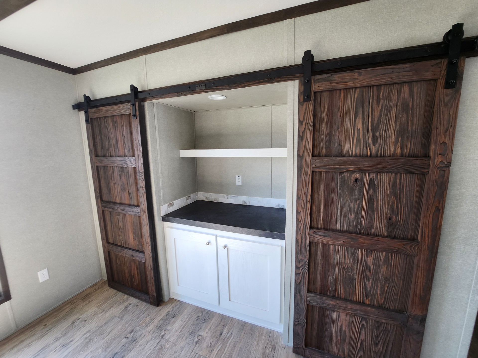 A room with rustic barn-style sliding wooden doors opens to a small alcove featuring white cabinets and a dark countertop. Light wood flooring enhances the cozy, farmhouse feel.