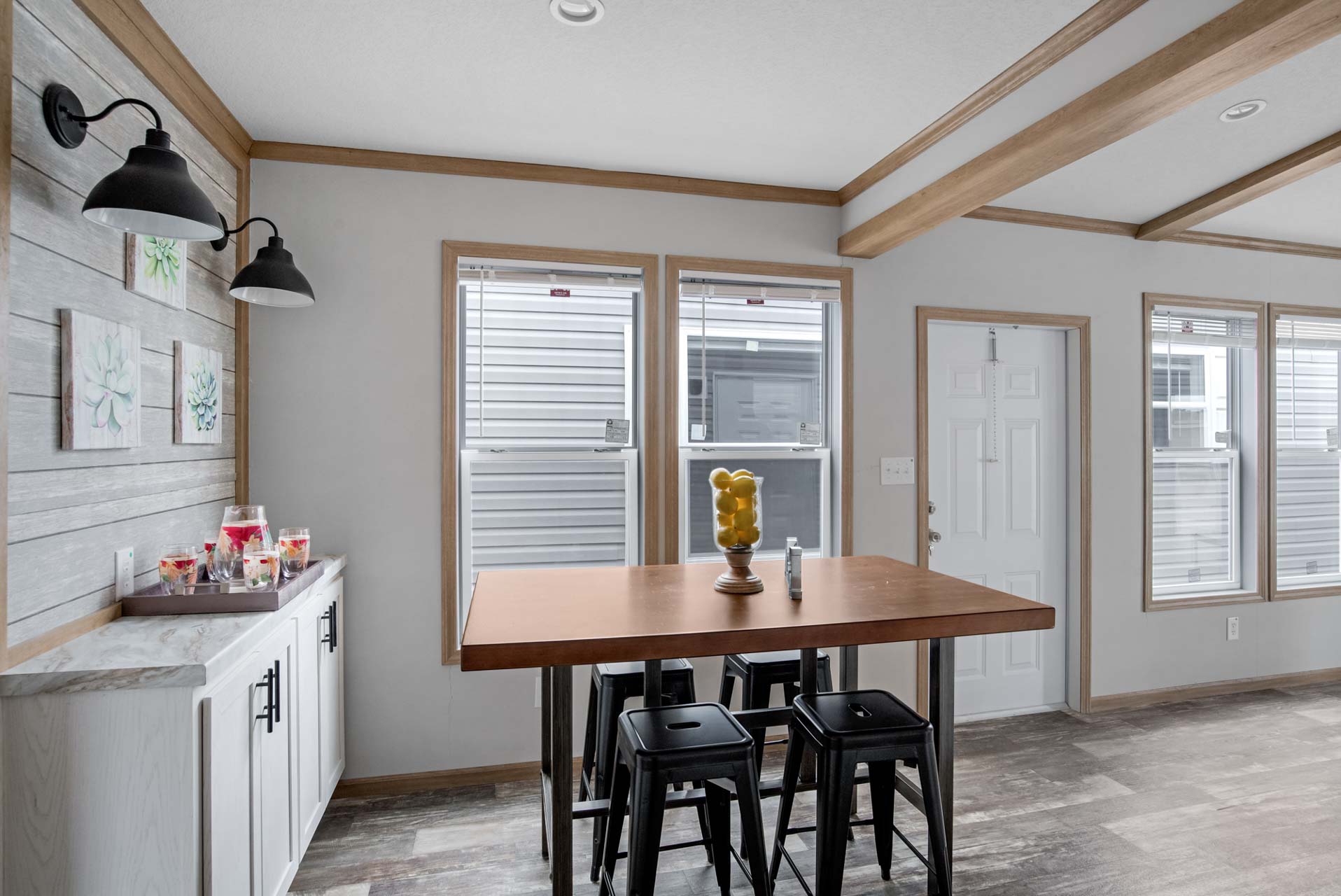 Modern dining area with a wooden table and black stools, a centerpiece of lemons. Left side features a white cabinet with drinks, under stylish wall lamps.