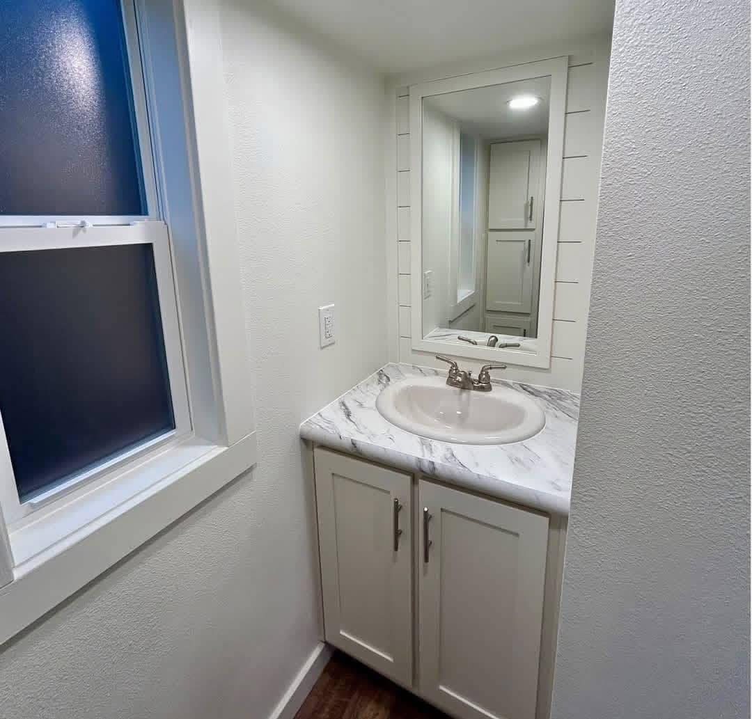 Small, well-lit bathroom with white walls. Features a marble countertop with a round sink, brushed metal faucet, and a large mirror. Frosted window on the left.