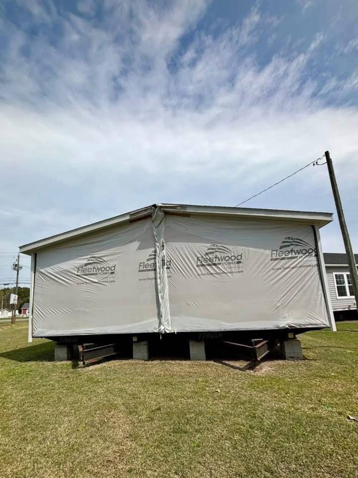 Large mobile home wrapped in white fabric with "Fleetwood" logos stands elevated on blocks in a grassy area under a partly cloudy sky.