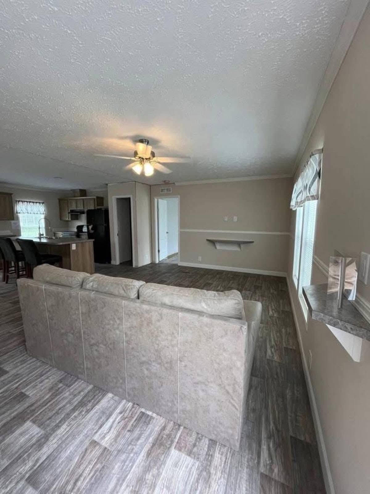 Spacious living area with light gray couch, wood-look flooring, ceiling fan, and neutral walls. Kitchen with bar stools visible in the background. Cozy and modern ambiance.