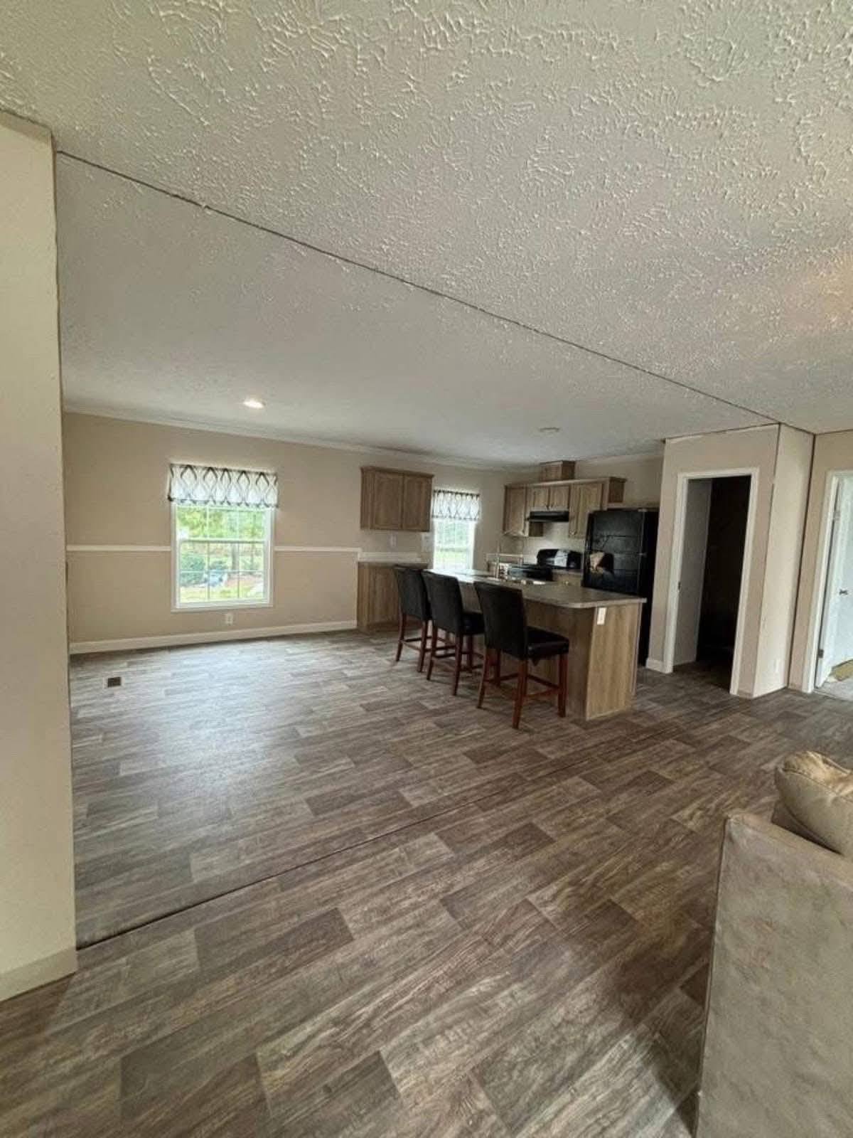Spacious open-plan kitchen and living area with wood-style flooring, neutral tones, wooden cabinets, and a breakfast bar with four dark chairs. Bright and inviting.