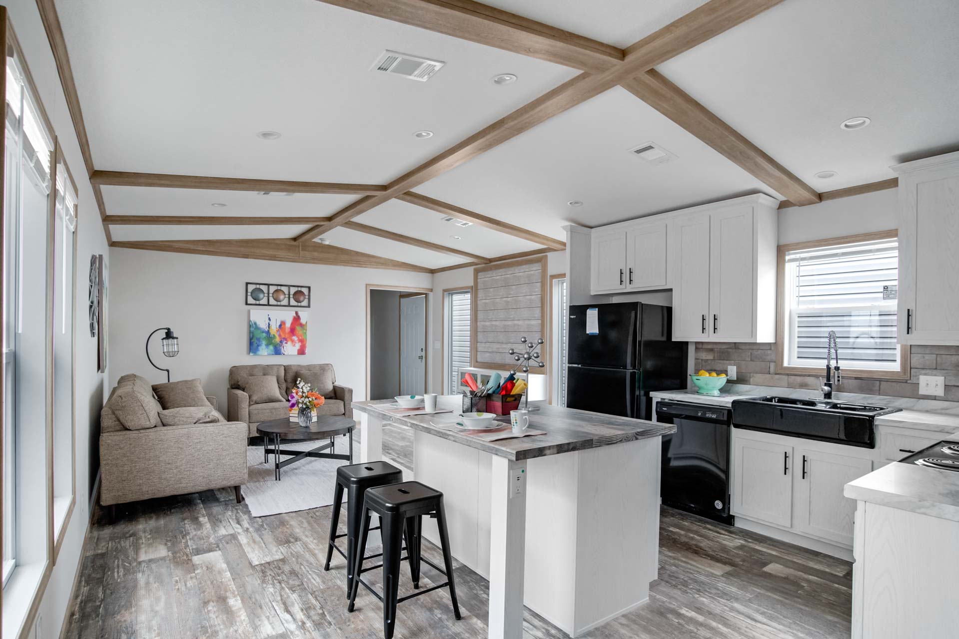 Modern kitchen and living area with light wood accents. Cozy beige sofa, colorful wall art, black appliances, and kitchen island with bar stools. Bright and inviting.