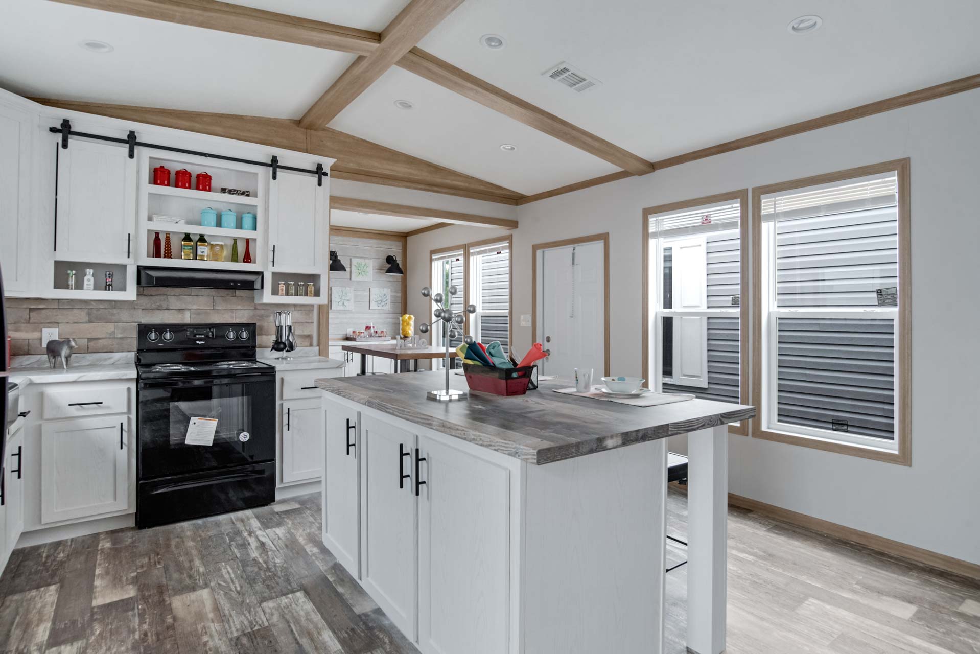 Bright kitchen with white cabinets, black appliances, and rustic wood details. Central island holds kitchen essentials. Windows bring in natural light.