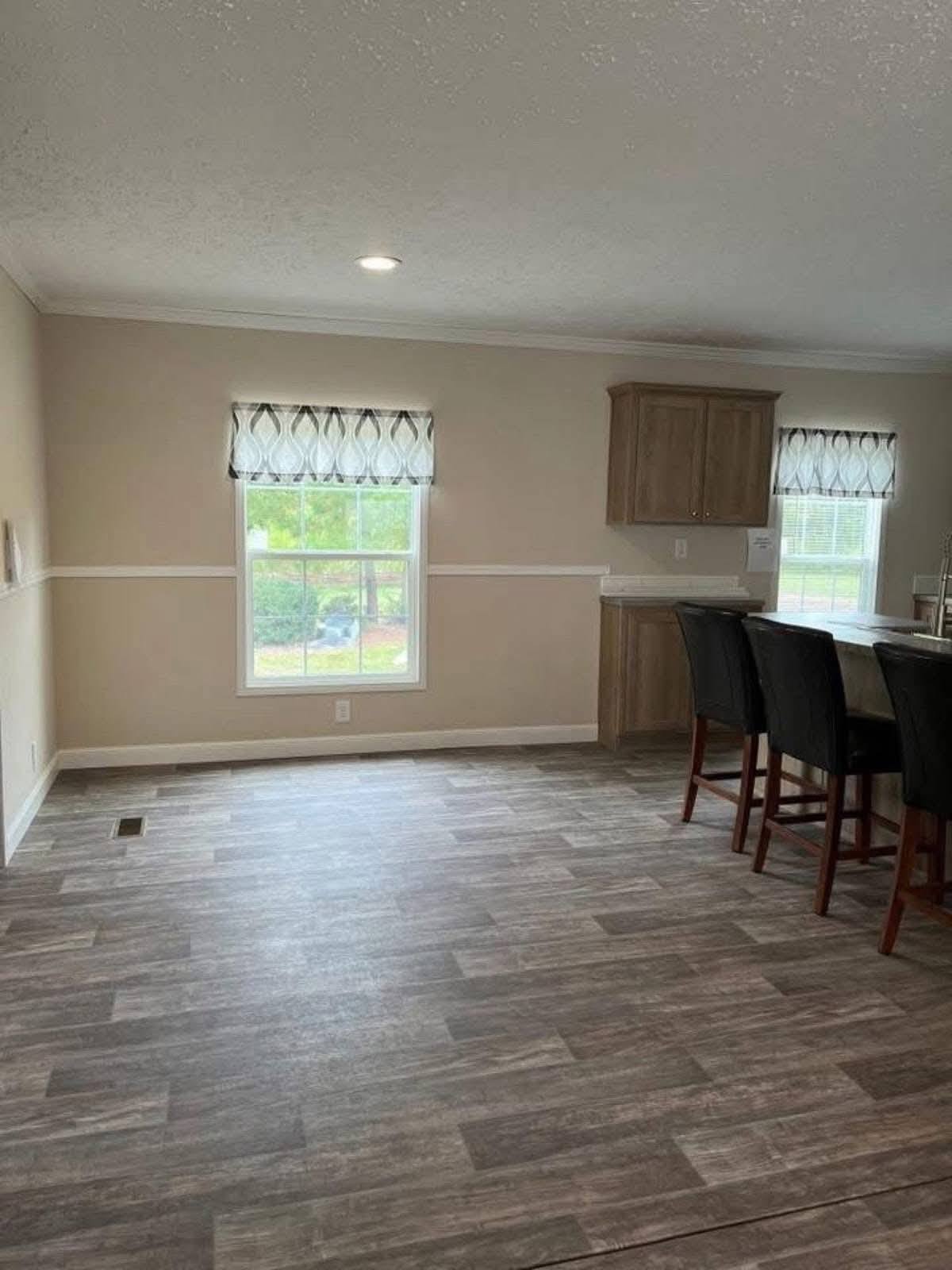A bright kitchen with light wood flooring, beige walls, and two large windows with patterned curtains. Three dark chairs line a countertop, creating a clean, open atmosphere.
