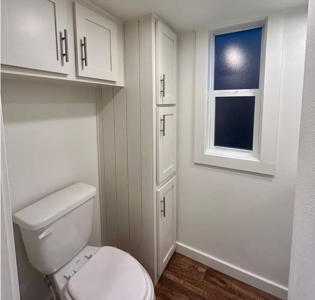 Compact bathroom with a white toilet, tall white storage cabinets, and a frosted window. Wooden floor creates a warm, clean, and bright atmosphere.