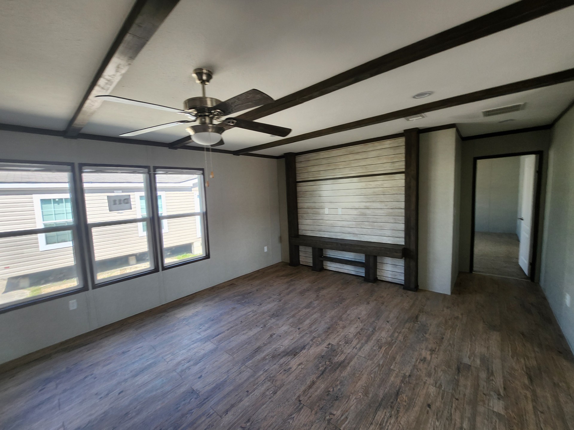 Spacious empty room with wooden floors and dark ceiling beams. Large windows on the left let in natural light. A ceiling fan and doorway to the right add to the airy feel.