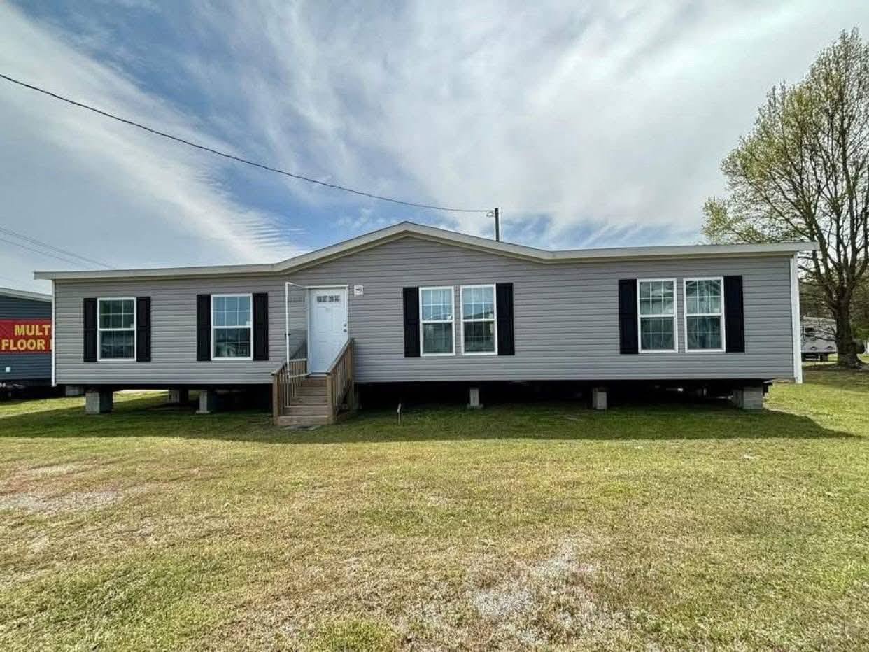 Single-story gray manufactured home with white trim, black shutters, and a front porch. Set on a grassy lot under a partly cloudy sky.