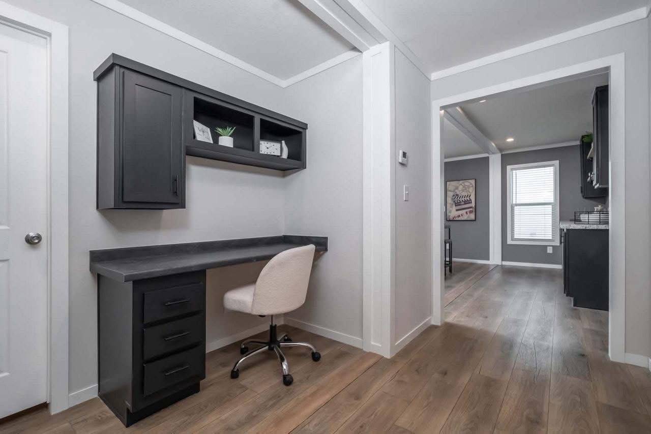 Minimalist home office nook with a gray desk, black cabinets, white swivel chair, and decor. Warm wood flooring leads to a sunlit, modern kitchen area.