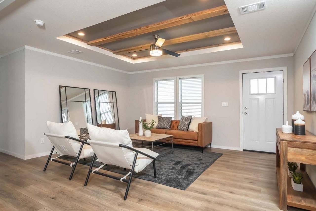Modern living room with tan sofa, white armchairs, and a black rug. Wooden ceiling beams and recessed lighting create a cozy, elegant atmosphere.