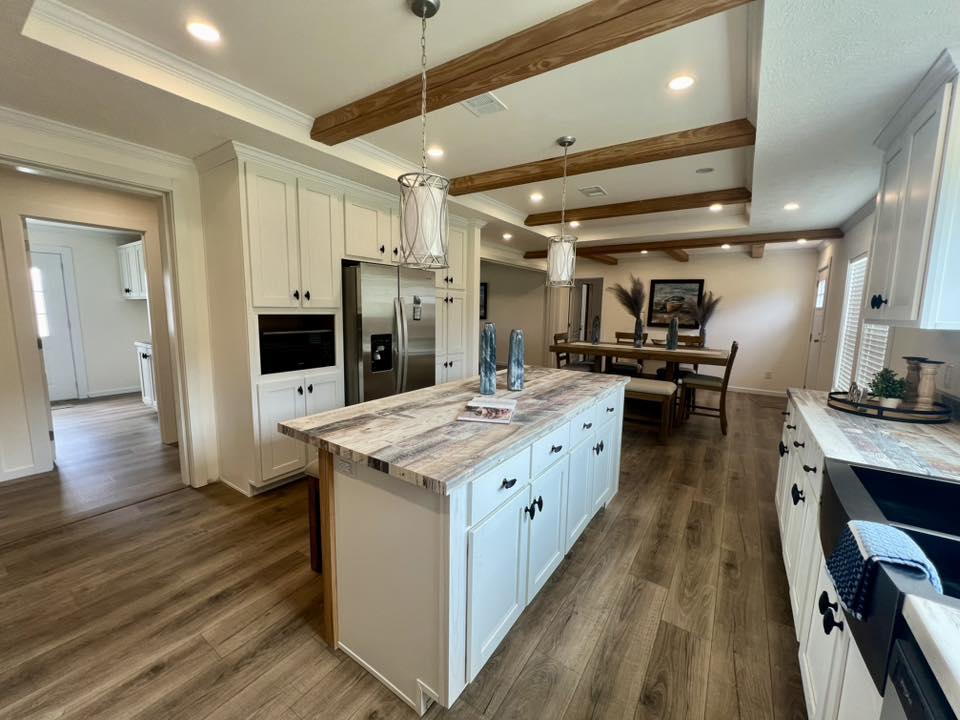 Spacious kitchen with a rustic feel, featuring wooden beams, white cabinets, large island, stainless steel appliances, and a dining area in the background.