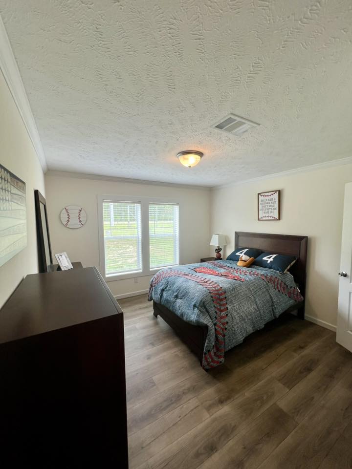 Cozy bedroom with wooden floor, twin bed with blue quilt, and numbered pillows. Baseball-themed decor on walls, soft natural light through a large window.