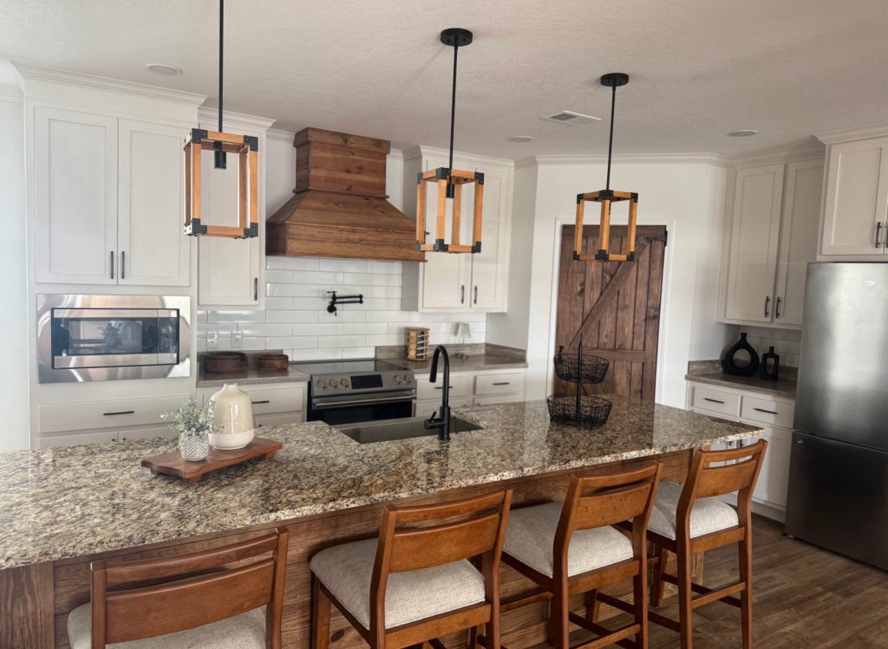 Warm, rustic kitchen with granite island, four wooden stools, wooden accents, and black fixtures. Soft lighting creates a cozy, inviting atmosphere.