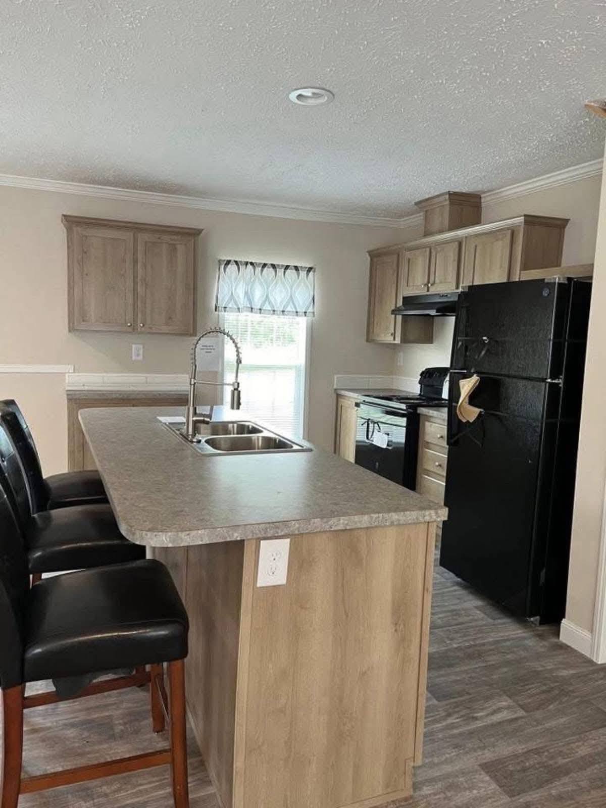 Modern kitchen with light wood cabinets, black appliances, and a central island with a stainless steel sink. Two dark chairs add contrast. Bright and tidy.