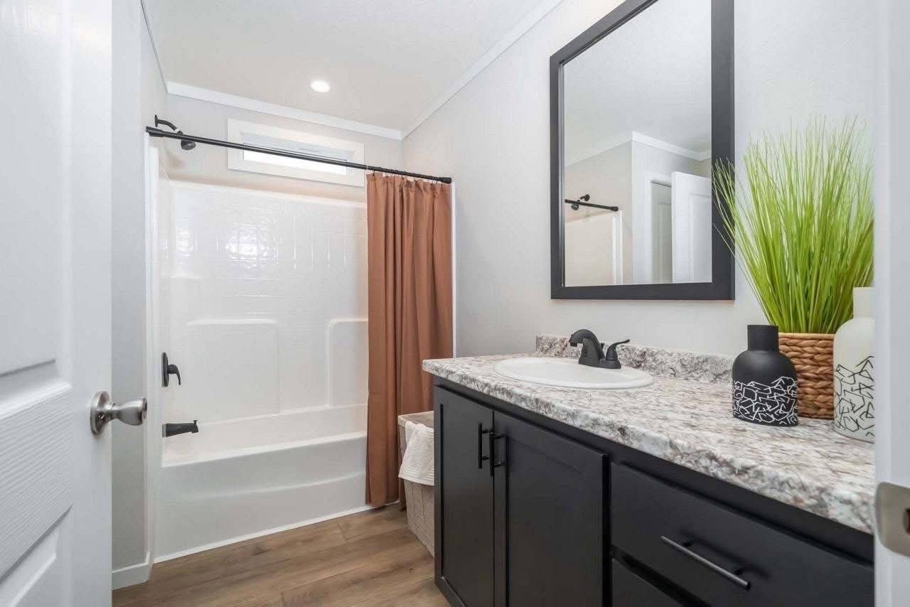 Modern bathroom with a white bathtub, brown curtain, and wood flooring. Granite countertop with black cabinets, a rectangular mirror, and decorative plants.