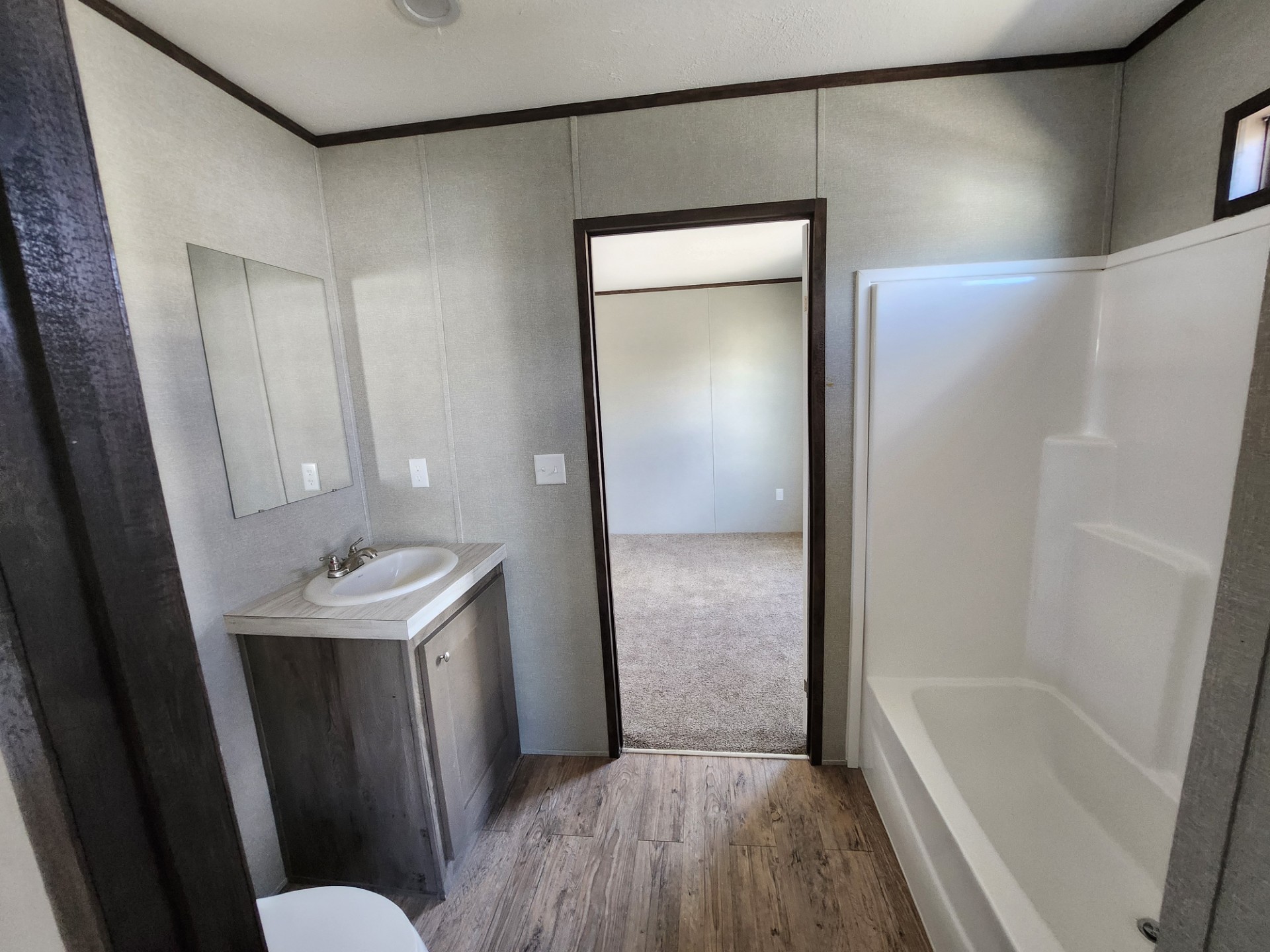 Small bathroom with neutral tones, featuring a wood vanity, mirror, and a white shower-tub combo. A door leads to a carpeted room. Cozy and clean atmosphere.