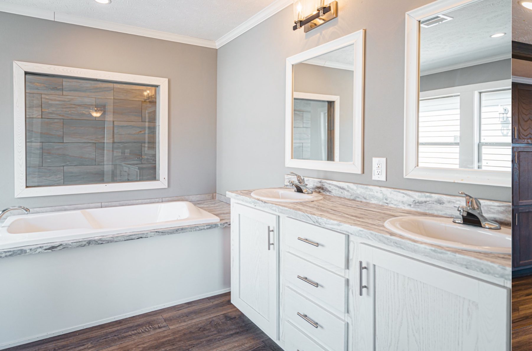 Modern bathroom with a gray double vanity featuring white cabinets and marble countertops. A large white bathtub sits beside a window with a wood tile accent, and the space has a bright, clean ambiance.