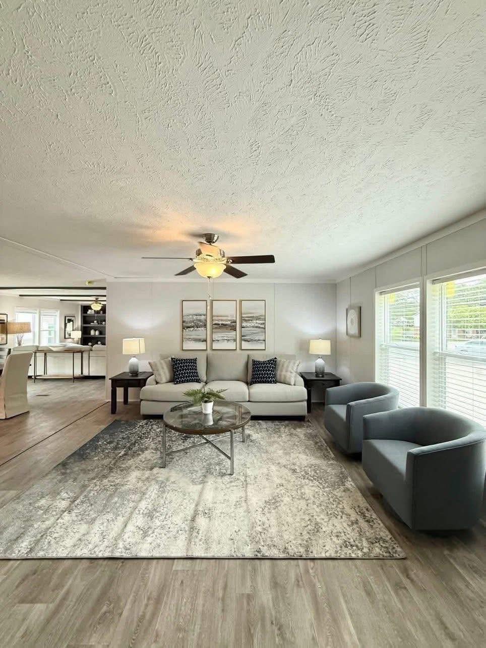 Elegant living room with a modern design. Features a white sofa, blue armchairs, abstract art, a ceiling fan, and a patterned rug, conveying a cozy, inviting atmosphere.