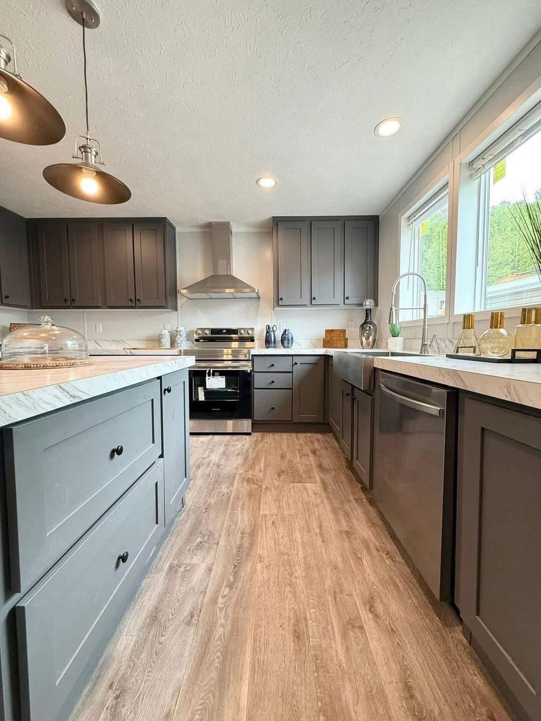 Modern kitchen with gray cabinetry, stainless steel appliances, and a light wood floor. Large windows provide natural light, creating a bright atmosphere.