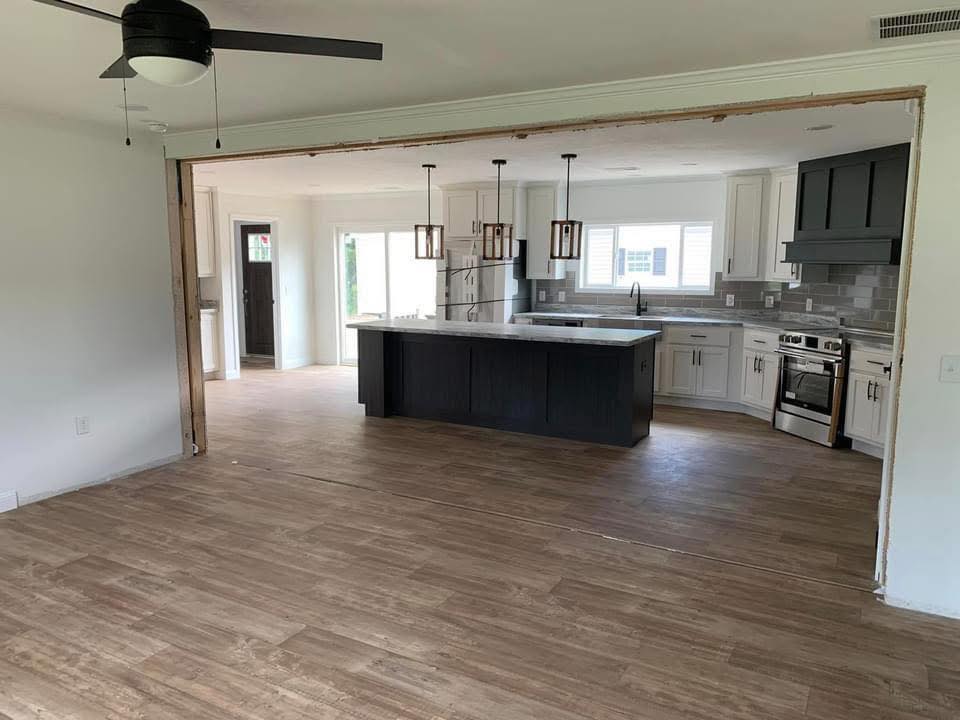 Open-concept room with brown wood flooring and a modern kitchen. Features a large island, stainless steel appliances, and pendant lights, creating a welcoming tone.