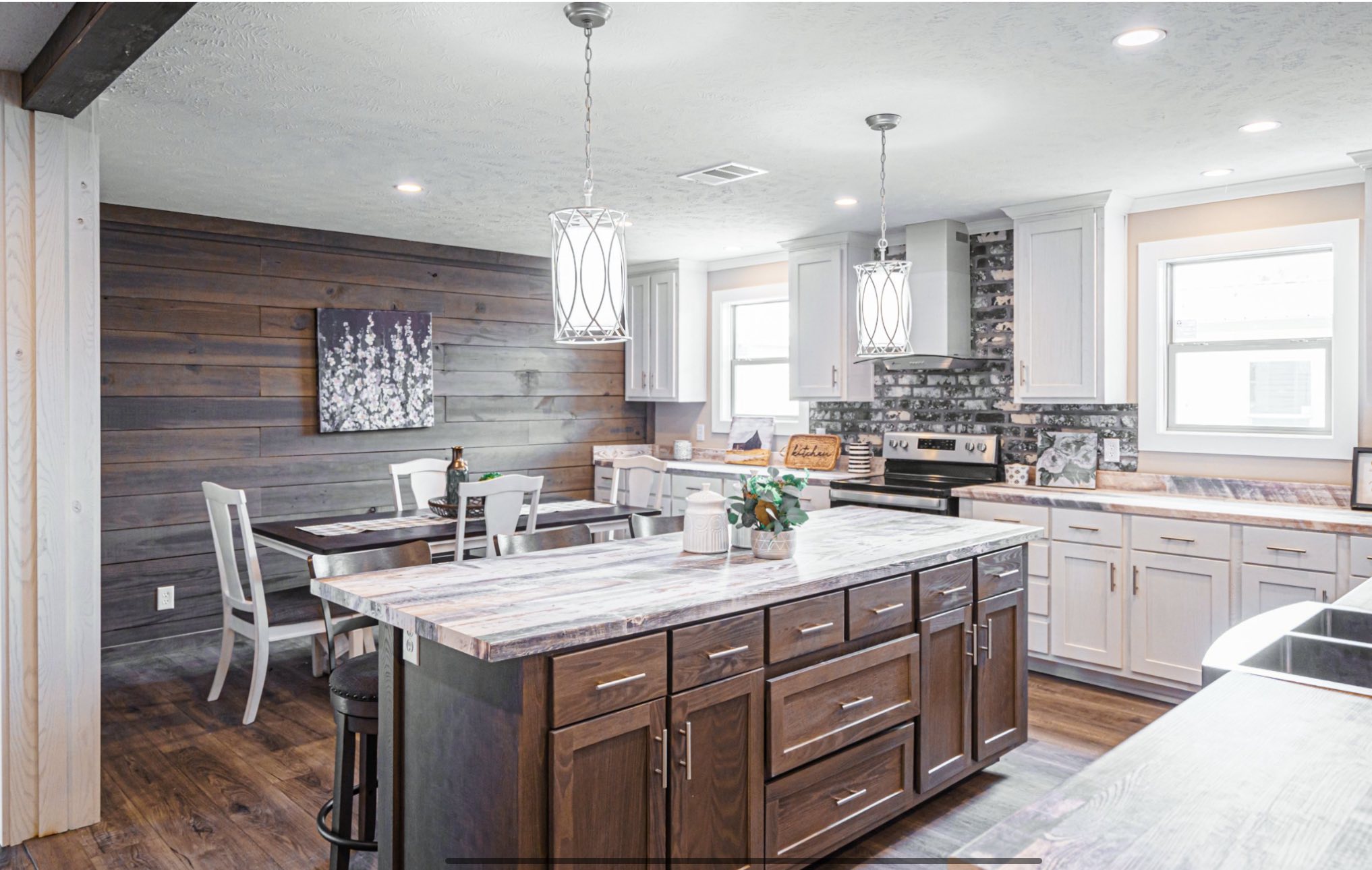 Modern farmhouse kitchen with a large island, marble countertops, and white cabinets. Two pendant lights hang above. Wooden accent wall adds warmth.
