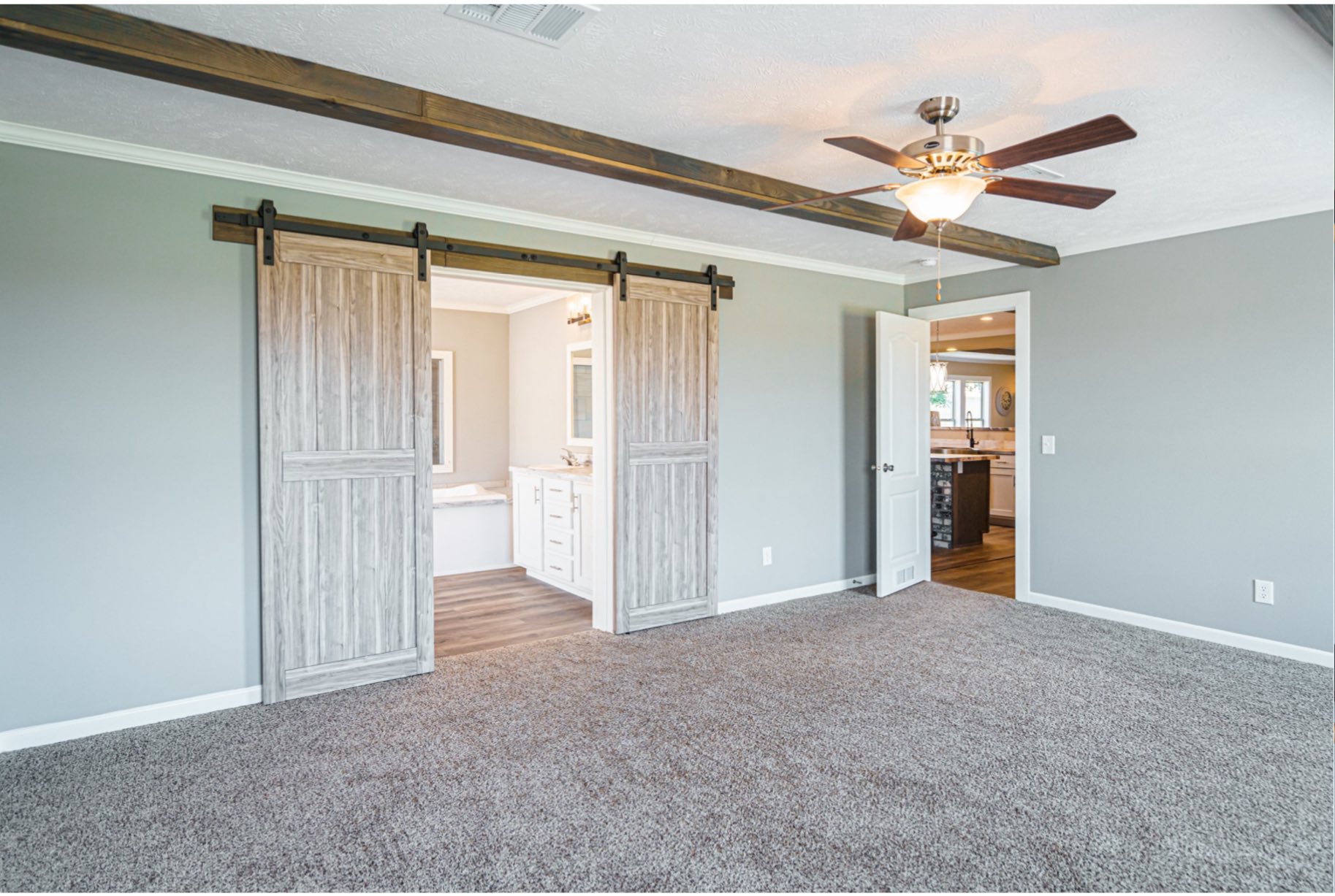 A spacious room with gray walls and carpet. Barn doors open to a modern bathroom. A ceiling fan hangs from a beamed ceiling, exuding warmth and style.