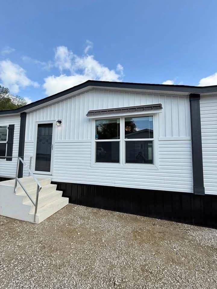 Modern white mobile home exterior with a gray door, large windows, and black shutters. Steps lead to the entrance. Clear blue sky backdrop.
