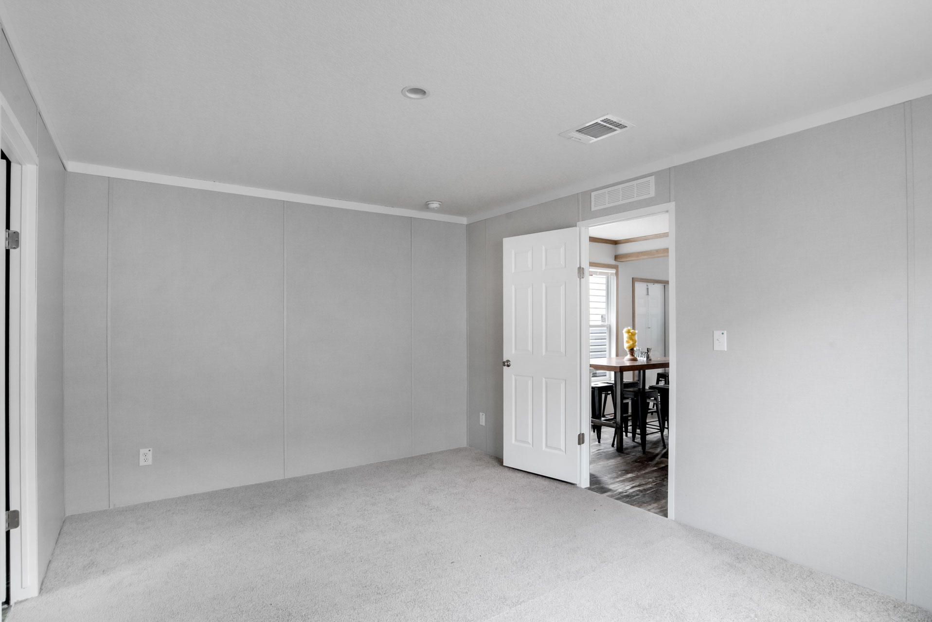Empty gray-walled room with a beige carpet and an open white door leading to a dining area. Bright, minimalist, and inviting atmosphere.