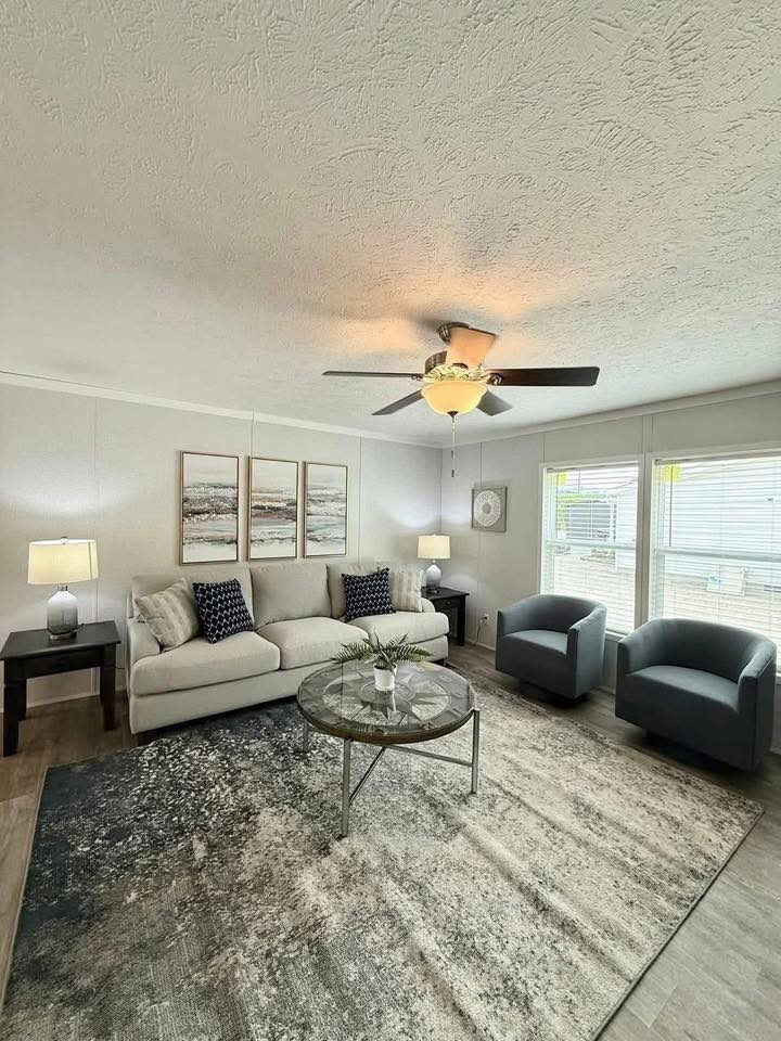 Bright living room with a beige sofa, two blue armchairs, and a round glass coffee table. Artwork and lamps add warmth. A ceiling fan is above.