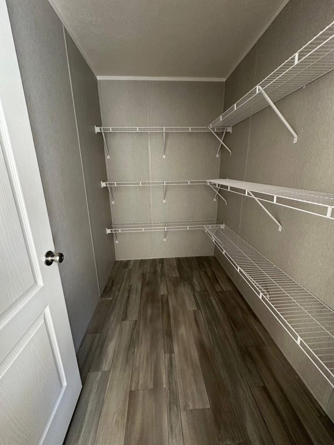Empty closet with gray walls and light wood flooring. Three tiers of white wire shelves line the right wall, offering ample storage space. The mood is minimalist and functional.