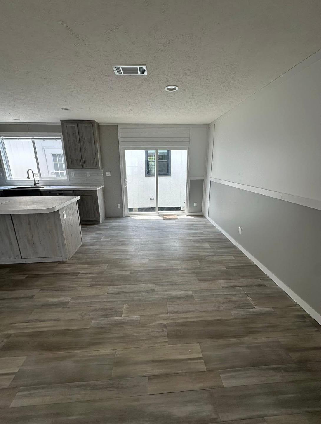 Spacious, empty room features hardwood floors, a kitchen island with gray cabinets, and sliding glass doors, allowing natural light to fill the space.