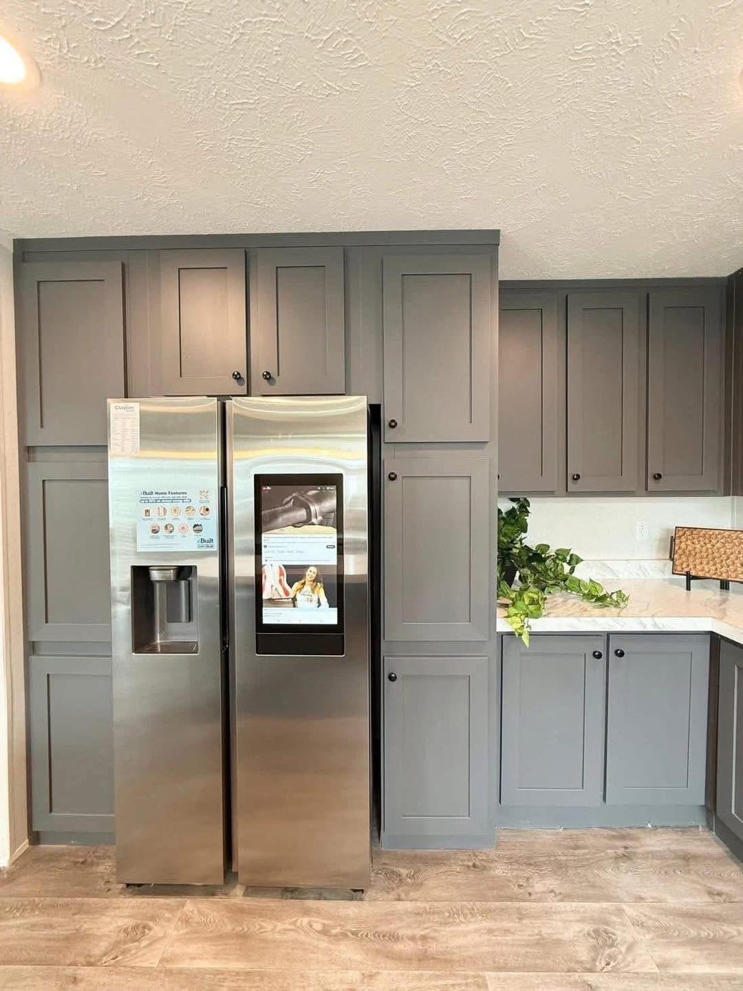 Modern kitchen with gray cabinets, a stainless steel fridge with a digital screen, and green plants on a white countertop. Light wood flooring.