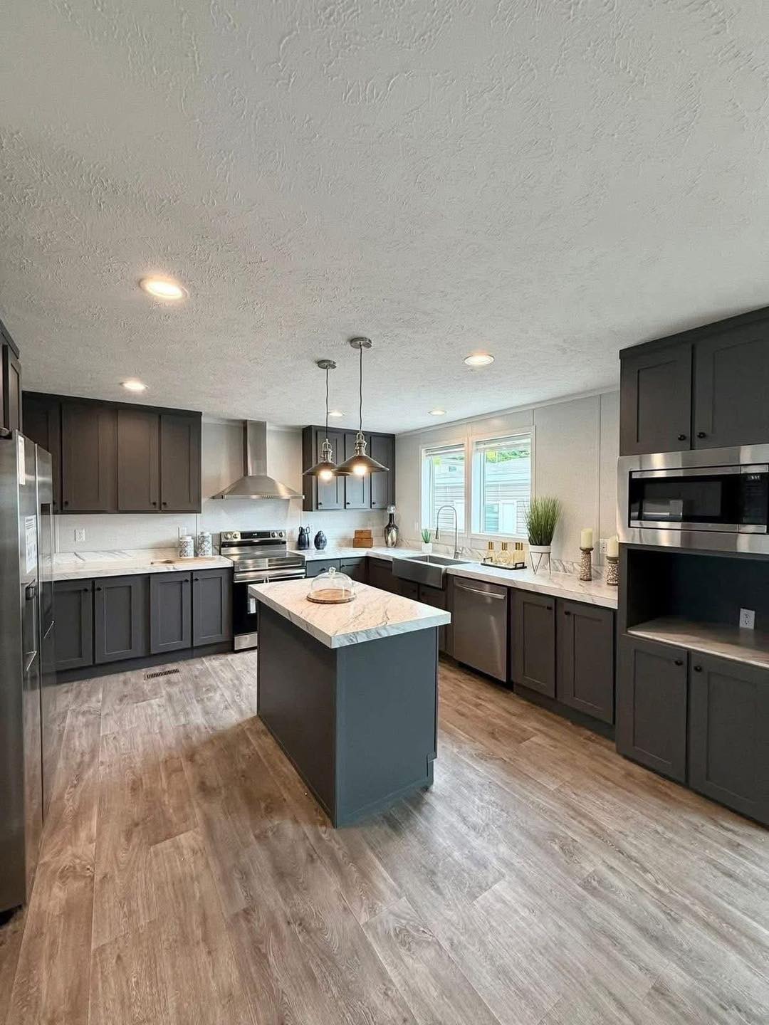 Modern kitchen with dark gray cabinets, a center island with marble countertop, wooden flooring, pendant lights, and bright natural light from windows.