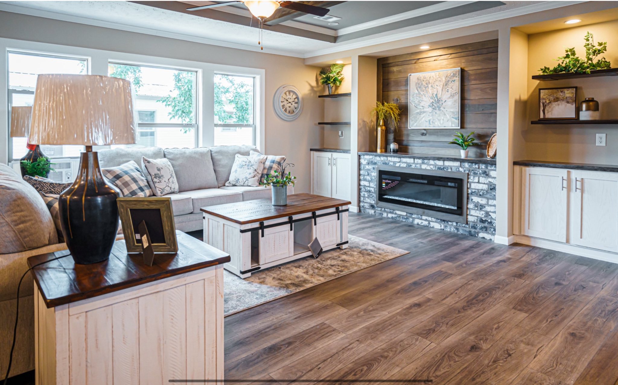 A cozy living room with a light gray sofa, plaid pillows, and a wooden coffee table. A fireplace is flanked by built-in shelves with plants. The room has warm lighting, wood floors, and a large window, creating a welcoming atmosphere.