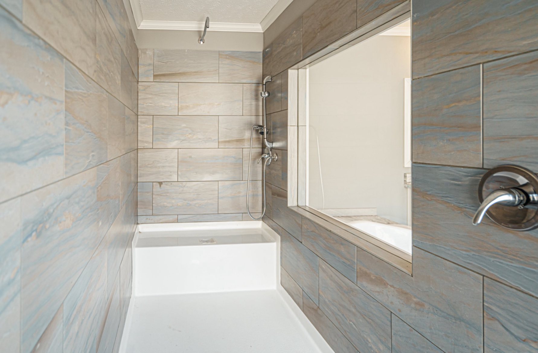 Spacious bathroom with earthy gray-blue tiled walls and a white bathtub. A large window opens to an adjacent room, creating an airy feel.
