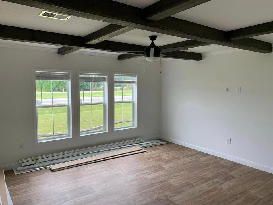 A bright, empty room with wood flooring. Three large windows reveal a grassy view outside. Dark ceiling beams and a fan add contrast and style.