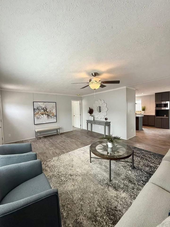 Modern living room with gray couches and a round glass coffee table atop a textured rug. A ceiling fan above and wall art add elegance to the cozy, open space.