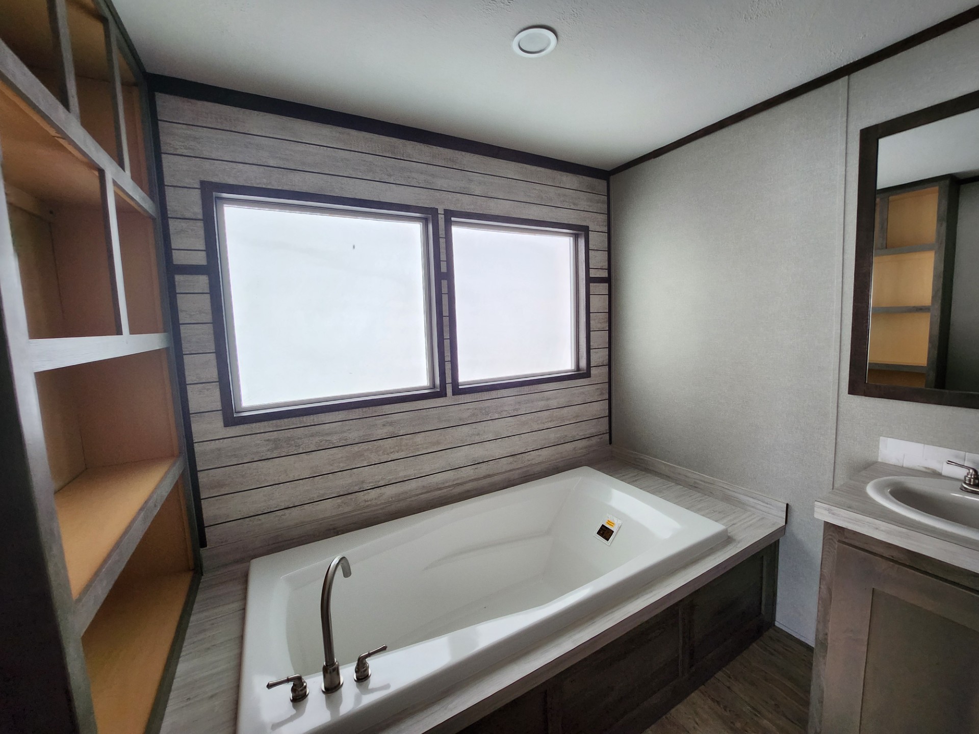 Modern bathroom with a large white tub beneath a window, framed by wooden panels. Open shelving to the left, a sink and mirror to the right.