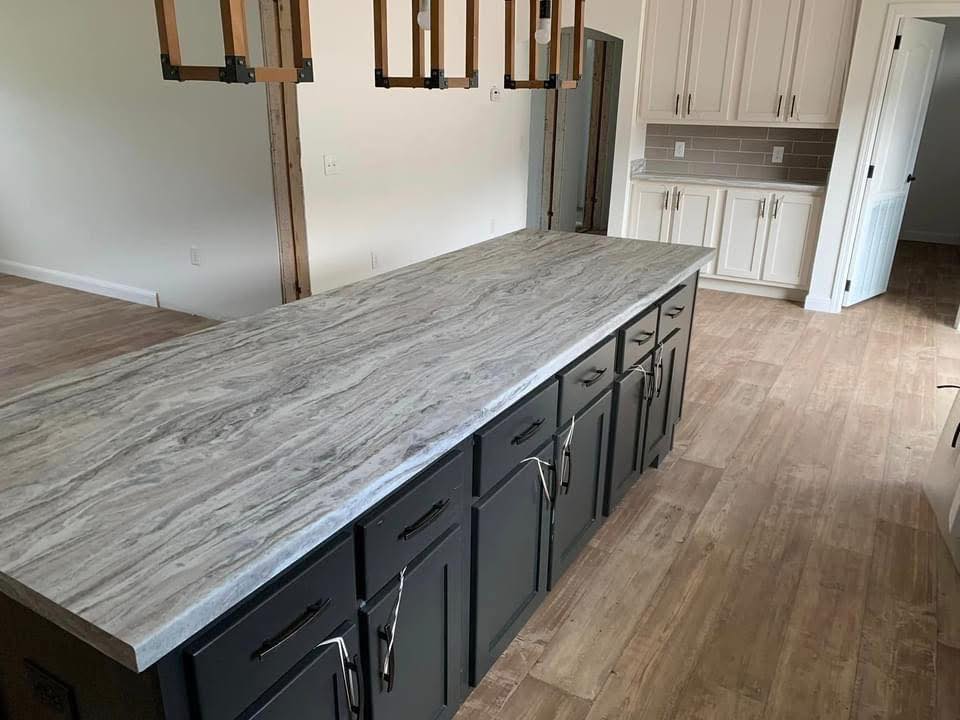 Spacious kitchen with a large marble island and dark cabinets, wooden floor, white cabinetry in the background, modern hanging lights, and neutral tones.