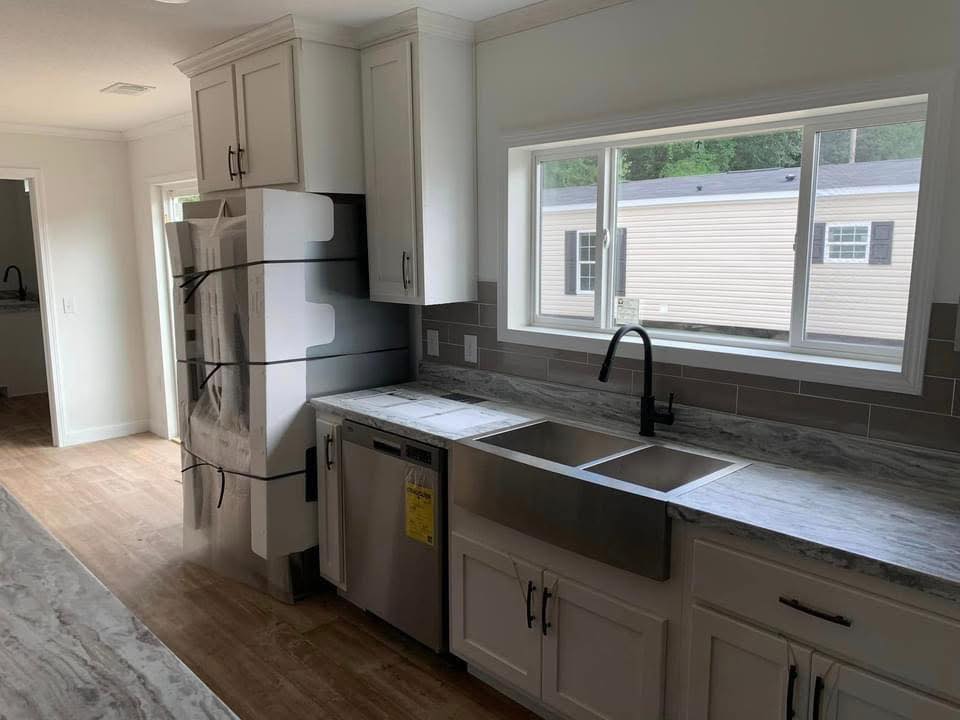 Modern kitchen with gray countertops, white cabinets, and a double sink below a large window. New appliances are wrapped in protective plastic.