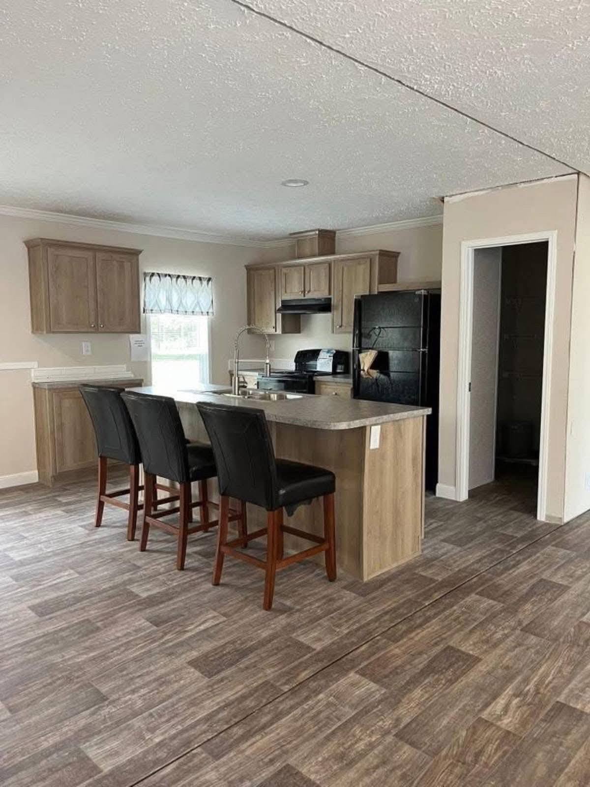 Modern kitchen with wooden cabinets and flooring, featuring a central island with three black chairs. Neutral tones create a cozy, welcoming atmosphere.