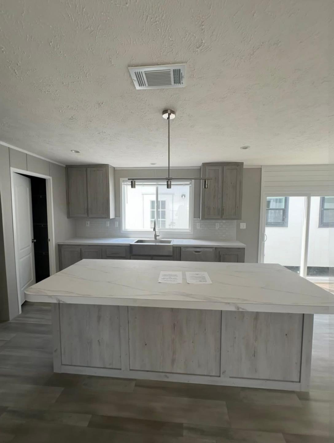 Modern kitchen with light gray cabinetry and a large marble island. Sunlit space features a central pendant light, plank flooring, and patio doors.