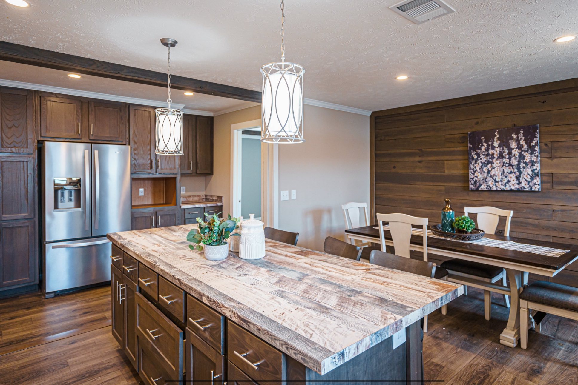Rustic kitchen with wood accents, featuring a large island with a textured countertop and hanging pendant lights. Stainless steel appliances and a dining table with a floral painting on the wooden wall create a warm, inviting atmosphere.